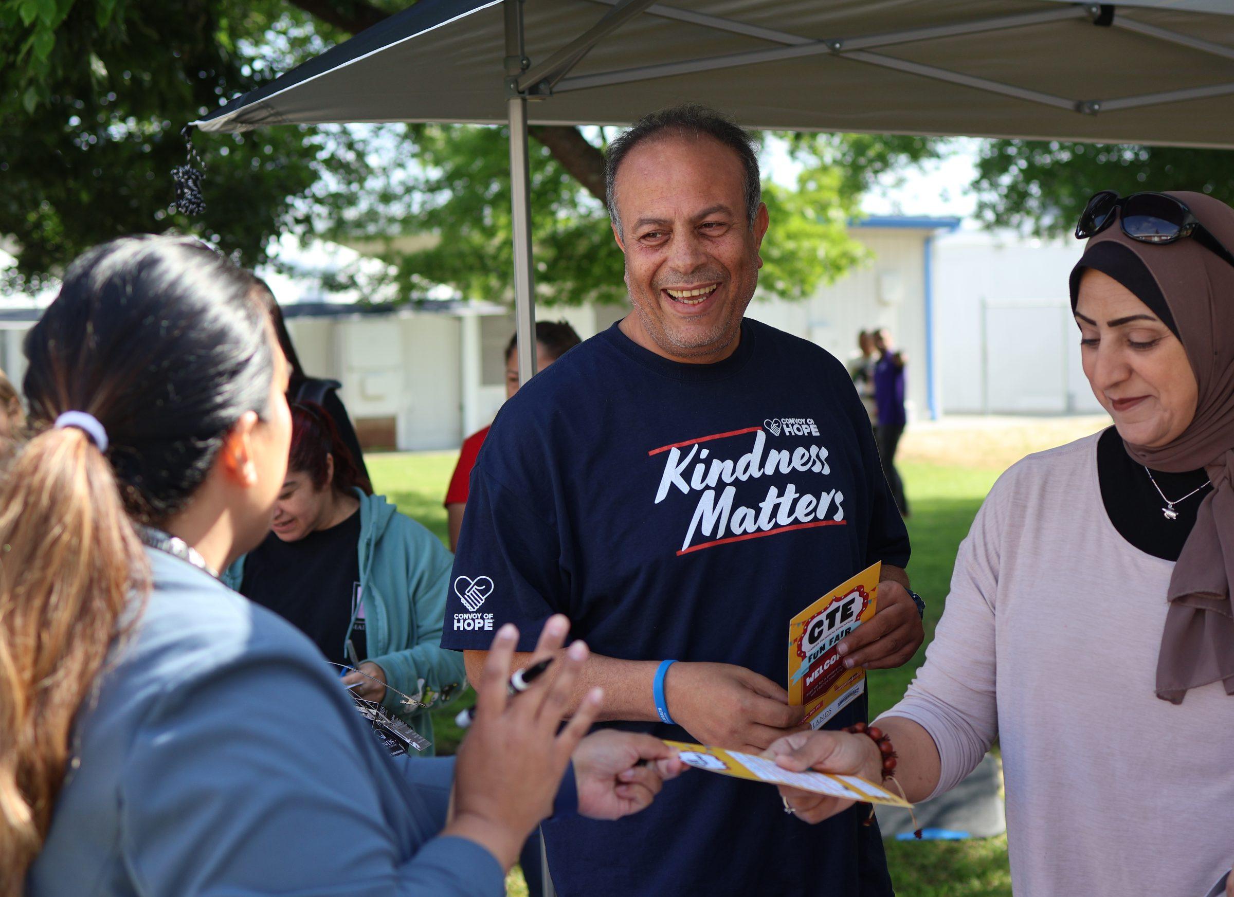A community partner sharing program information with two community members under an outdoor tent.
