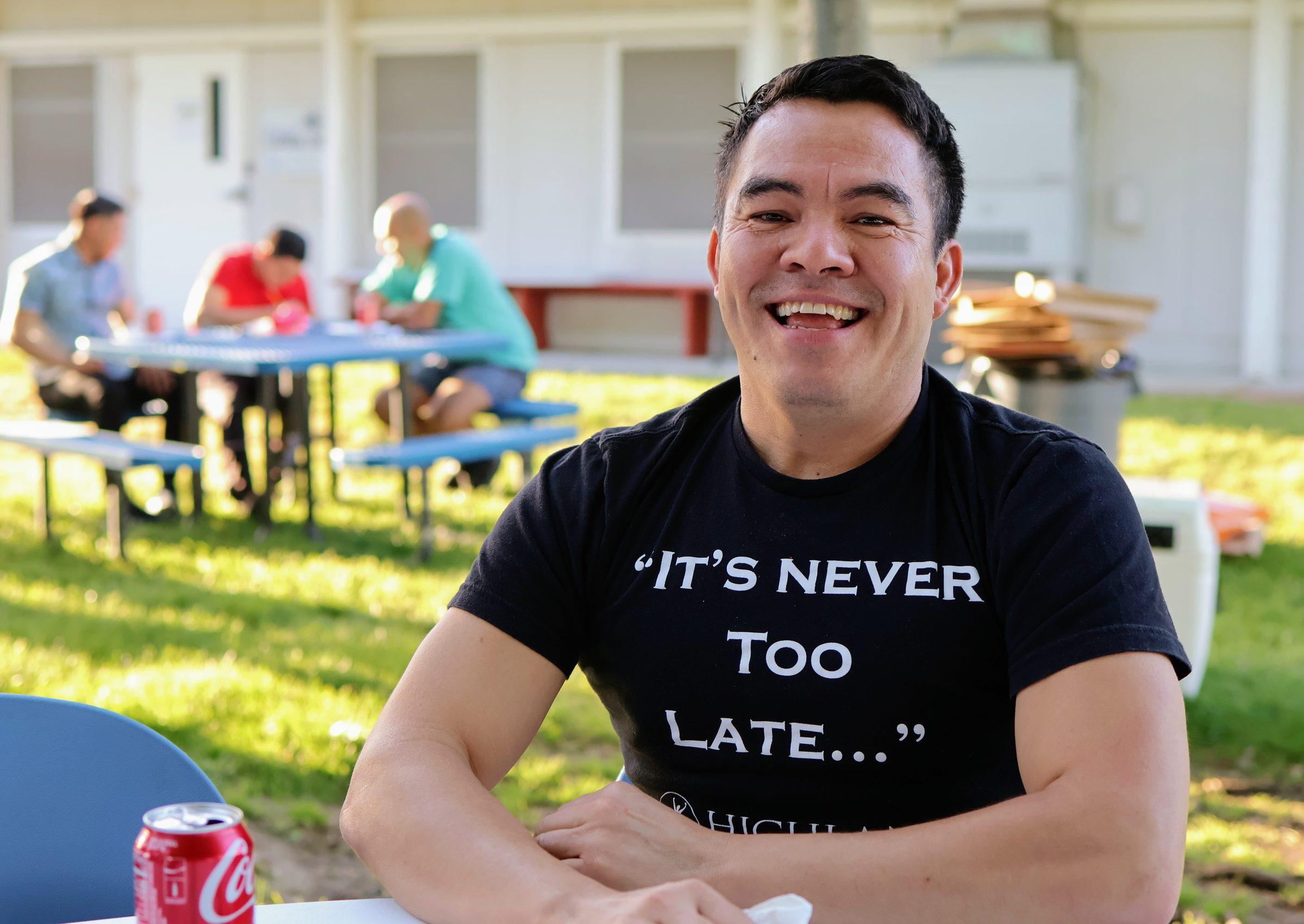 A smiling HCCTS community member wearing a shirt that reads "It's Never Too Late..." at an outdoor community gathering.